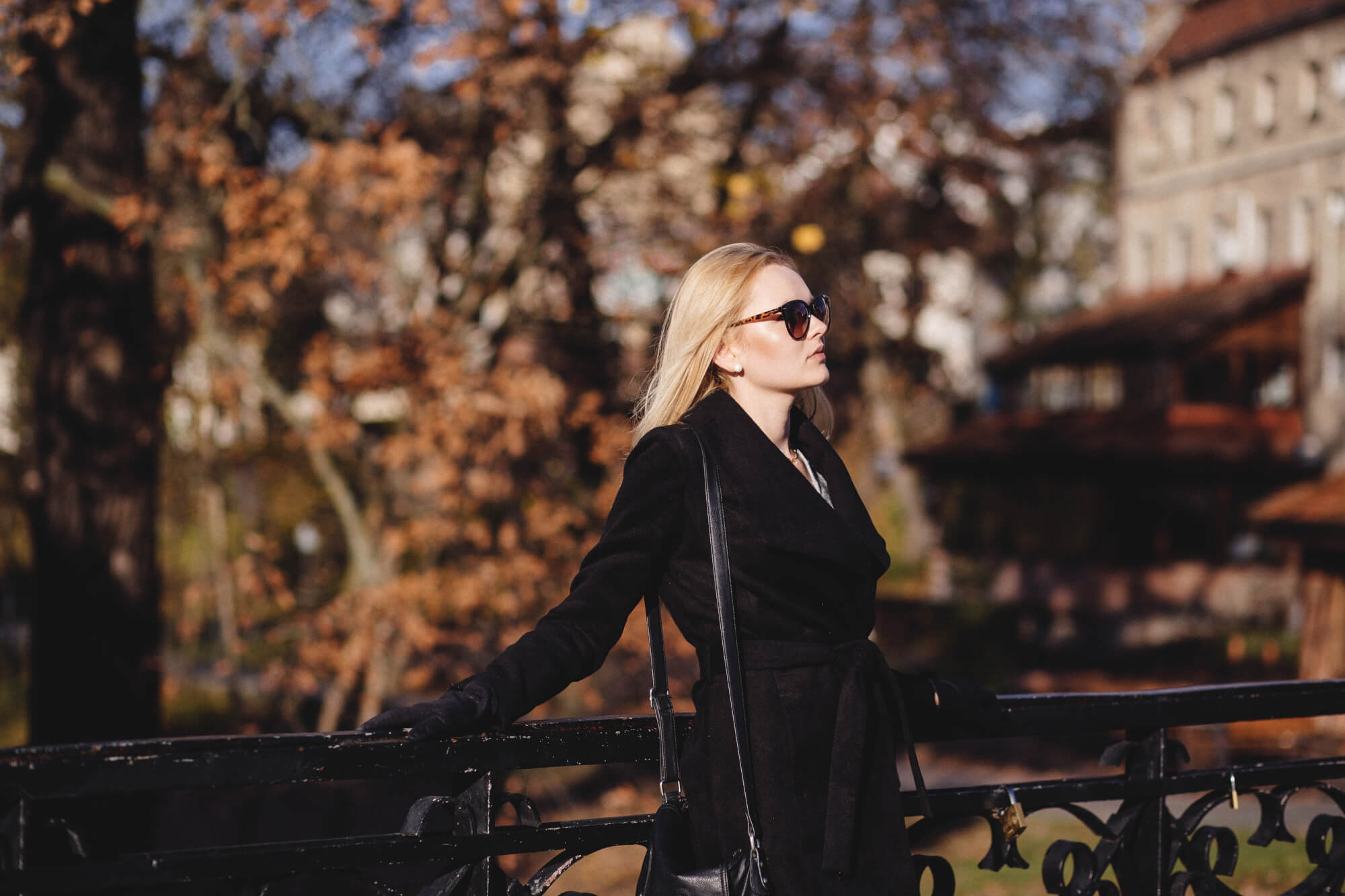 Glamorous looking model with blonde hair and sunglasses on standing against an iron railing with autumn trees out of focus in the background.