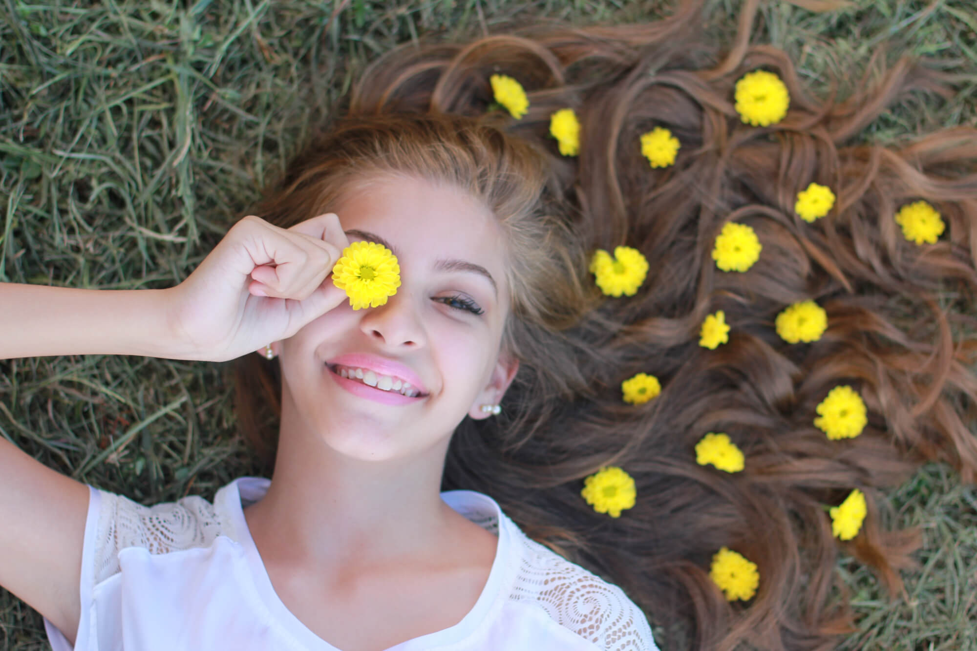 A young female model lying on the grass, smiling with yellow flowers in her hair whilst holding one of the flowers over her right eye.