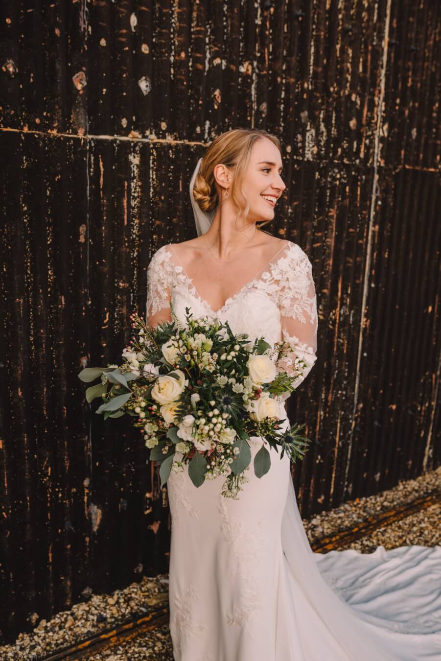 Bride smiling and looking away from the camera whilst holding flowers. Bridal hair and makeup by Bristol makeup artist Evie Smith