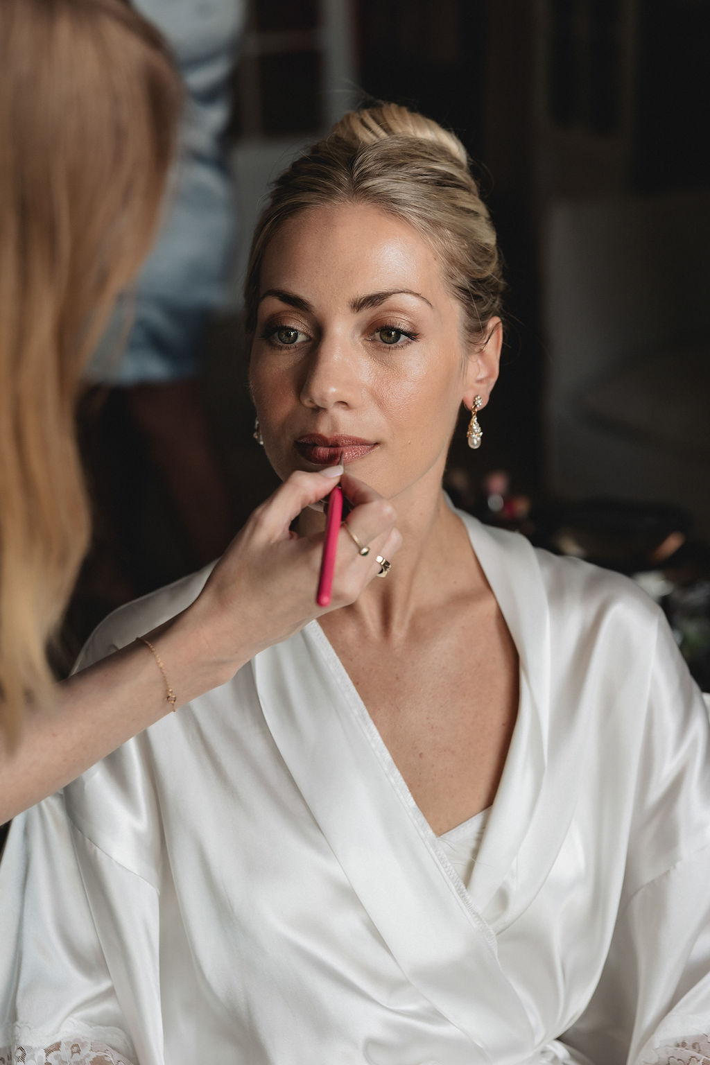 A beautiful bride sat in a white dressing gown having her lipstick applied on her wedding day.