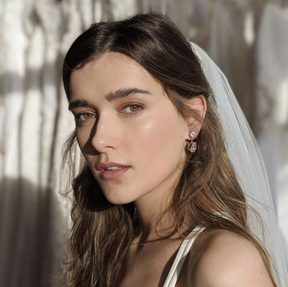 A brunette woman wearing a veil and a white strappy wedding dress. she is looking directly into the camera, showing her brown eyes and bridal wedding makeup