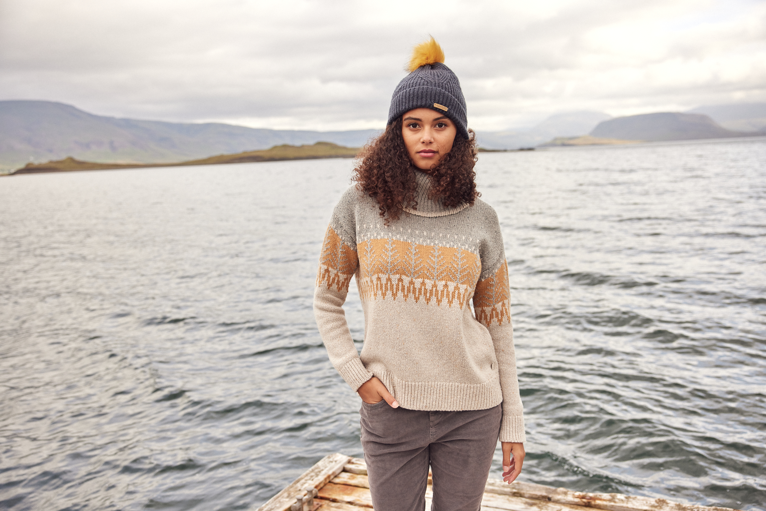 Female model in jumper and bobble hat standing in front of a lake in Iceland whilst shooting for Weird Fish. Makeup provided by Bristol Makeup Artist Evie Smith.