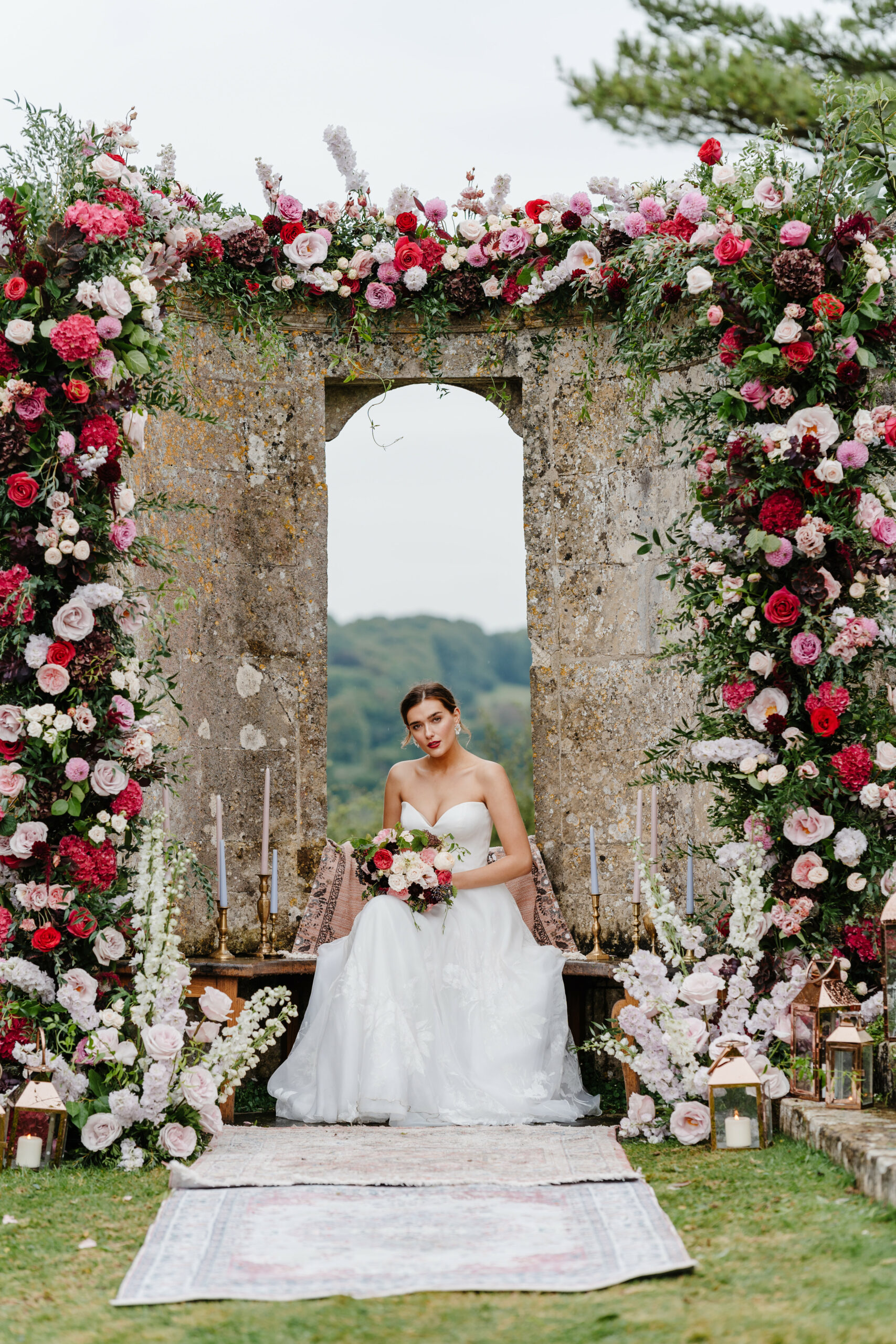 A bride sitting in a stone archway in the rolling hills of the Cotswolds. She is wearing a white wedding dress and holding flowers. she is surrounded by flowers and wearing red lipstick