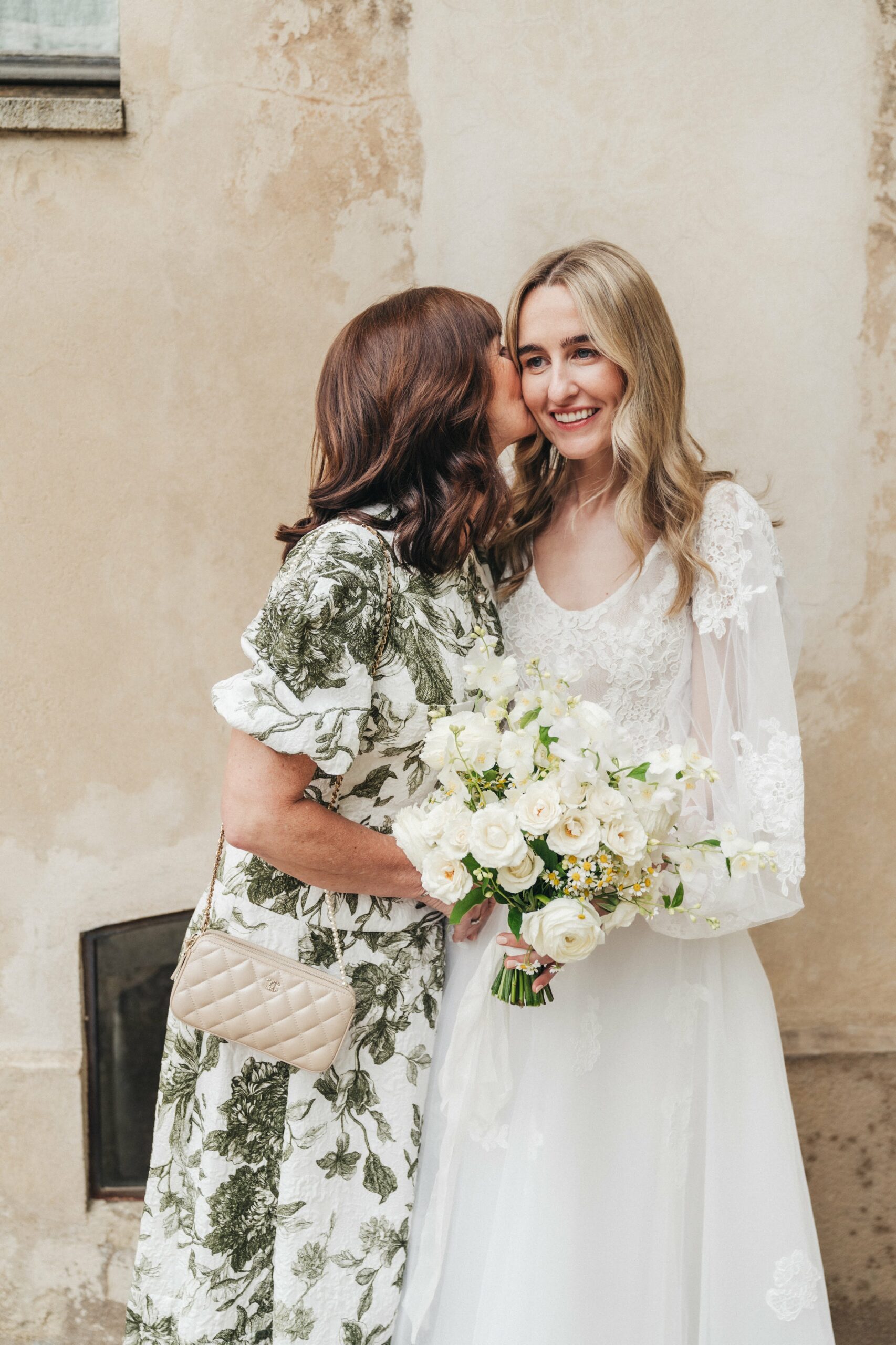 Smiling bride in a flowing white wedding dress and radiant makeup shares a moment with a close friend or family member.