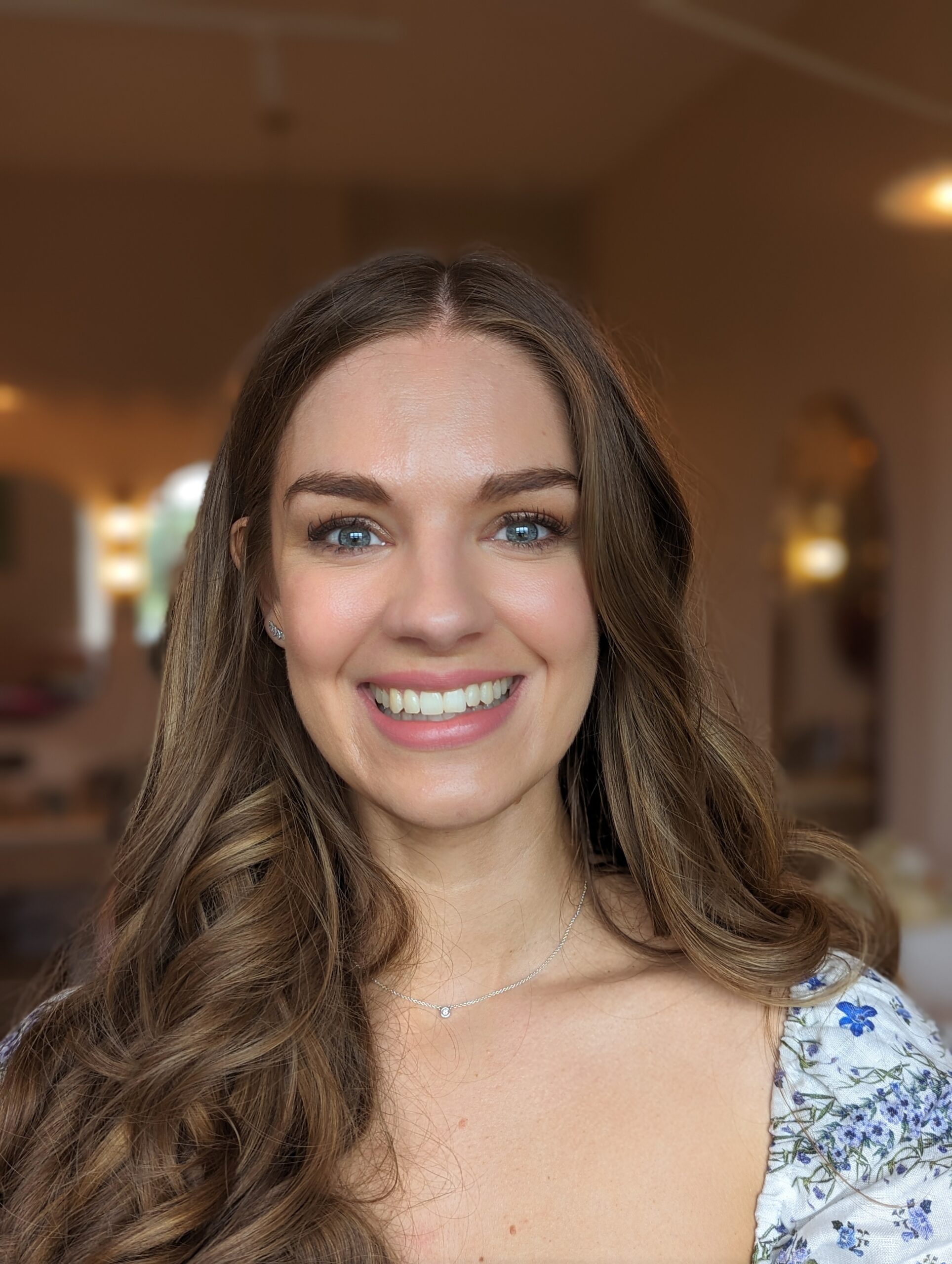 brunette woman staring into the camera smiling. she is wearing a blue floral dress and her hair is brushed over one shoulder