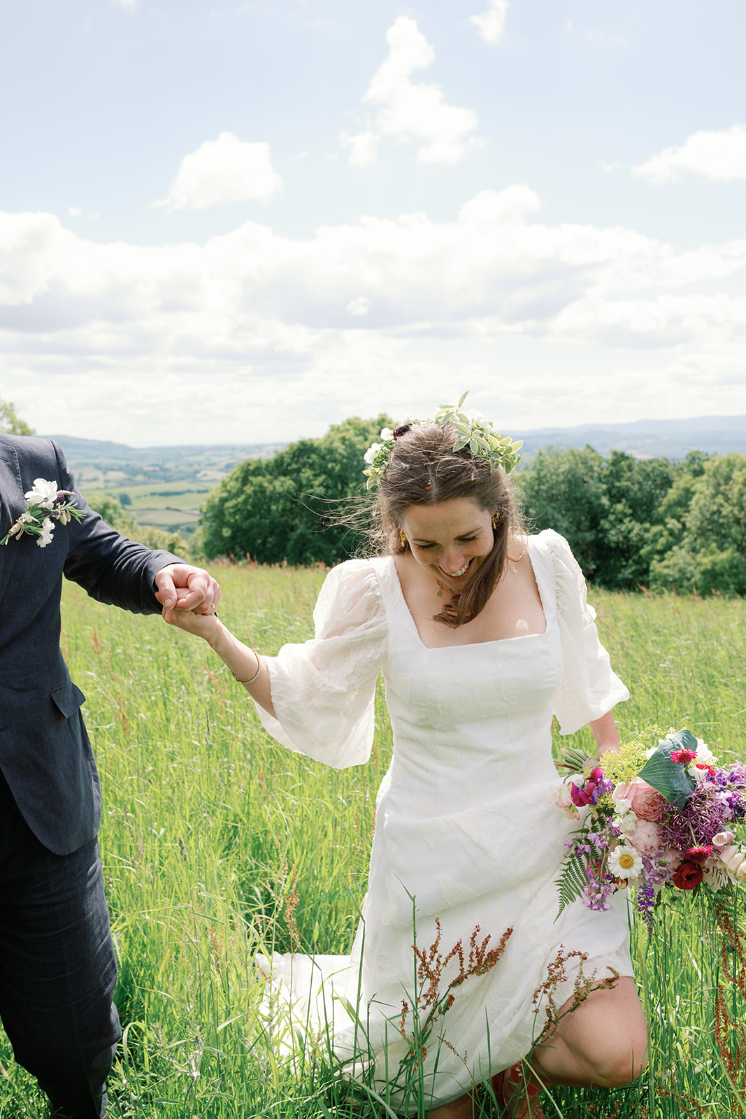 A bride and groom walking through a flower meadow on their wedding day. The bride has flowers in her hair and is looking down whilst holding a bouquet of flowers