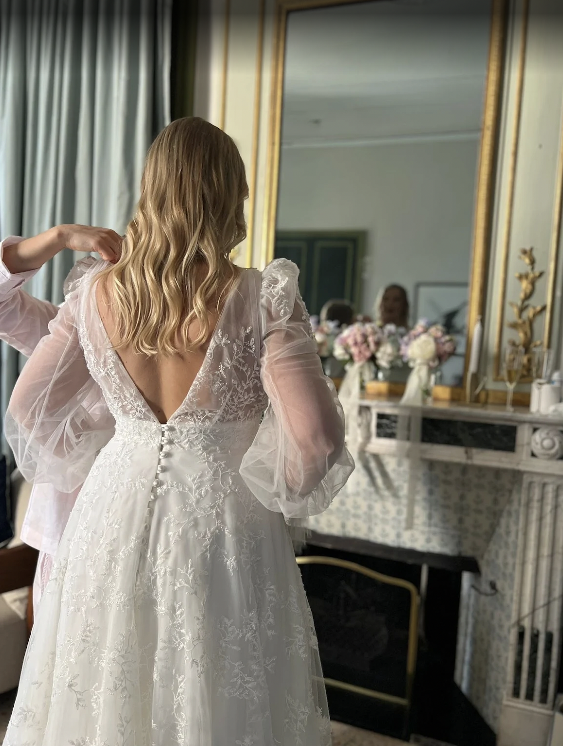 a bridal with soft waves in her hair looking into a mirror wearing a white wedding dress.