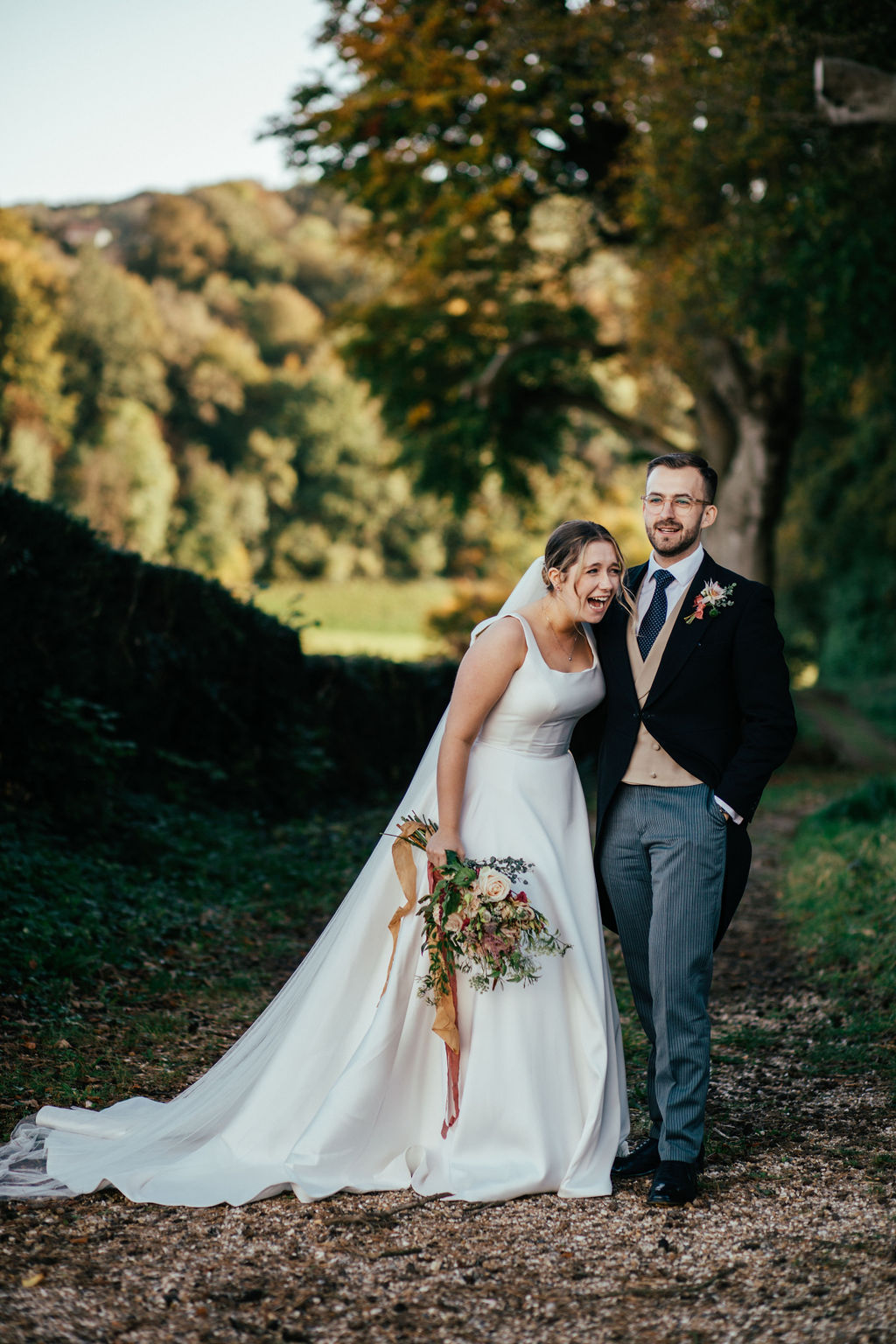 a bride and groom laughing in the countryside. the bride is holding a bunch of flowers with orange ribbon and laughing.